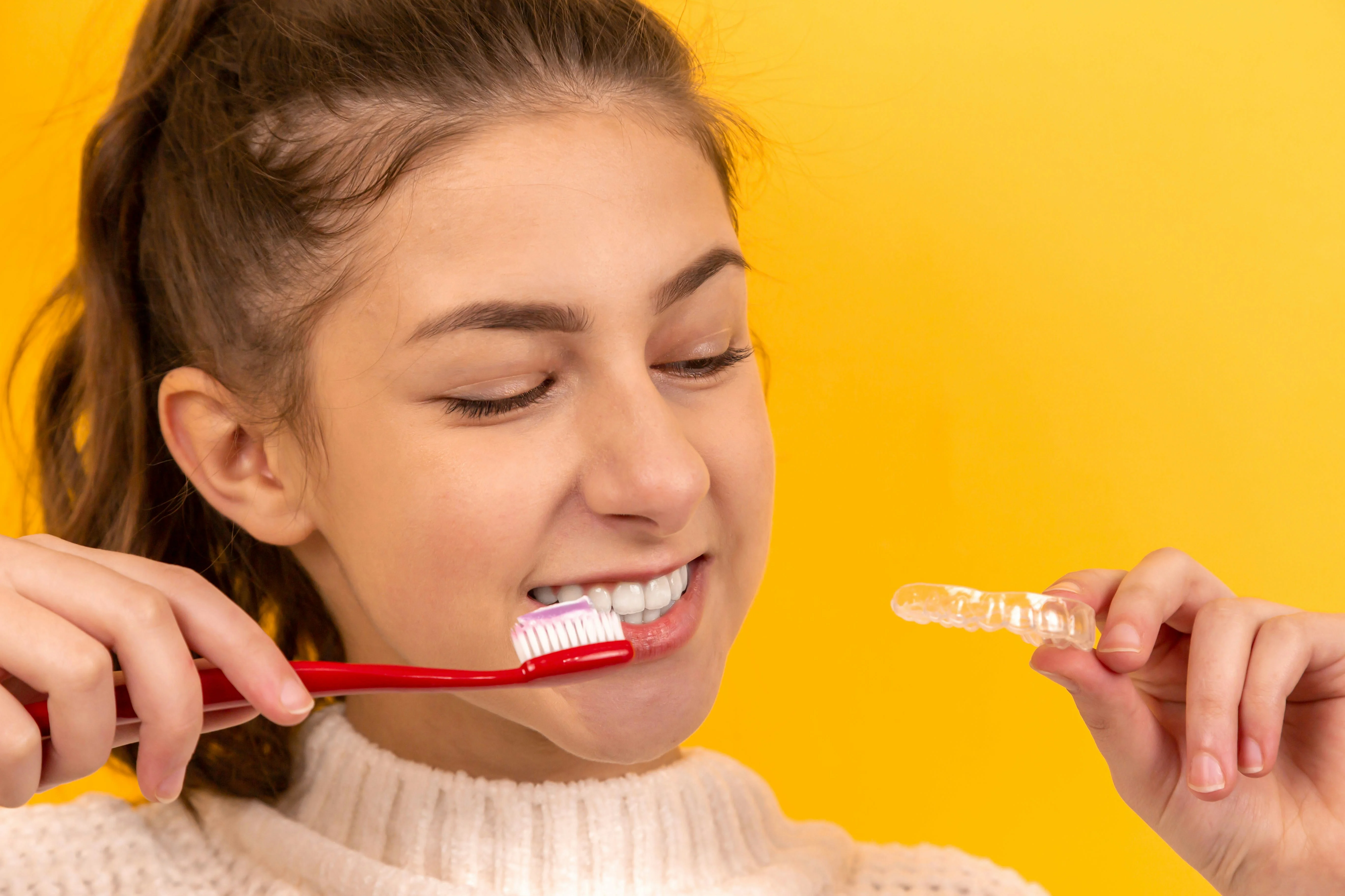 Smiling teenage girl brushing teeth while holding clear aligner against yellow background.